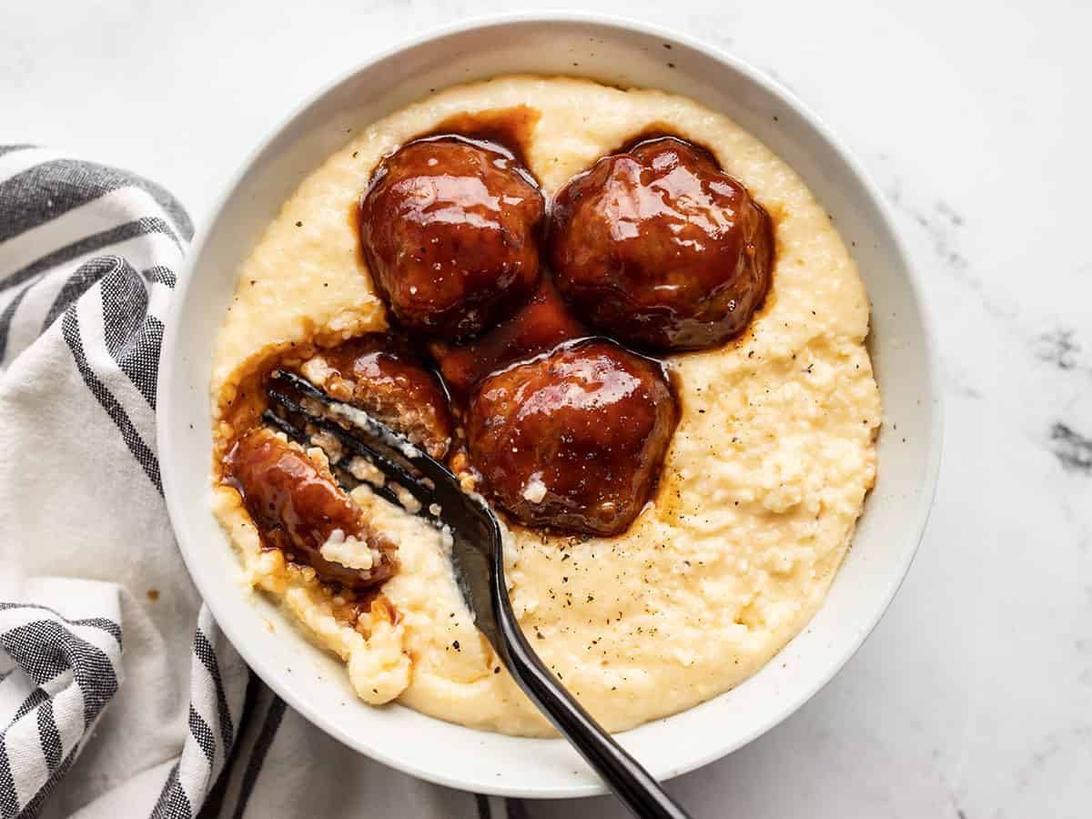 A fork cutting into a bbq meatball on a bed of cheese grits