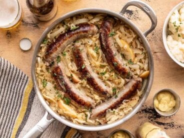 Bratwurst and sauerkraut in a skillet with beer, potatoes, and mustard on the sides