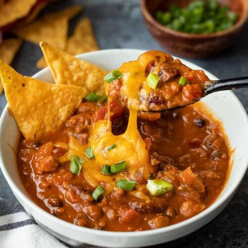A spoon lifting a bite of pumpkin chili with melted cheese stretching from the bowl