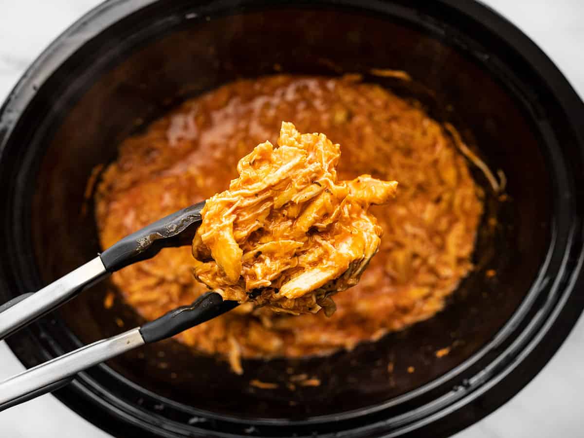 tongs holding a scoop of buffalo chicken over the slow cooker