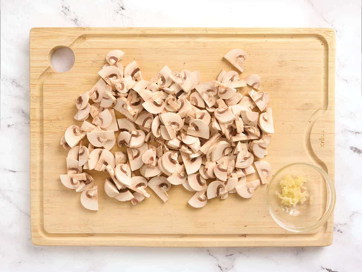 Sliced mushrooms on a a wooden chopping board with minced garlic in a bowl.