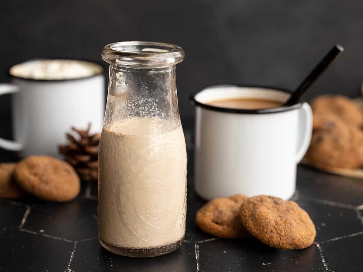 gingerbread coffee creamer in a glass milk bottle with coffee cups and cookies in the background