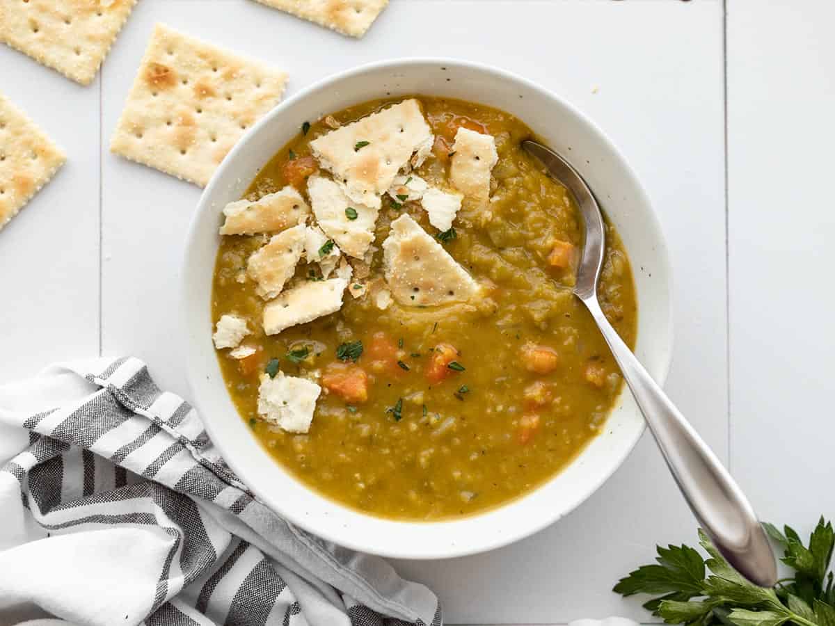 A bowl full of split pea soup with saltine crackers, a spoon in the side