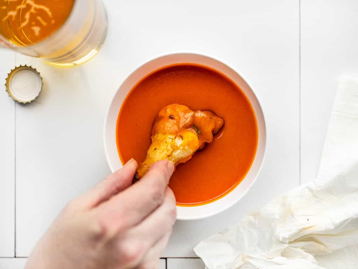 a hand dipping a chicken wing into a bowl of hot sauce