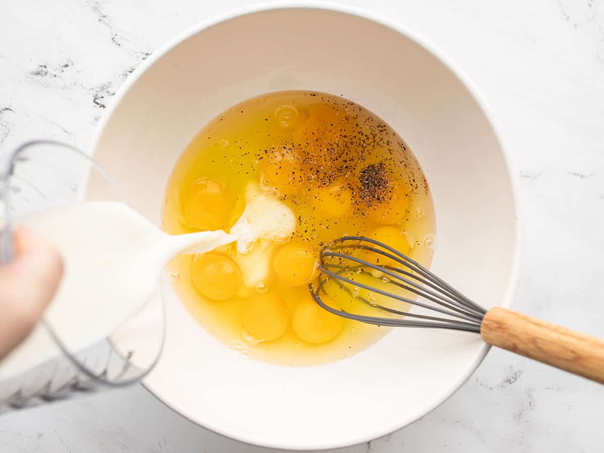 milk being poured into a bowl of eggs with a whisk