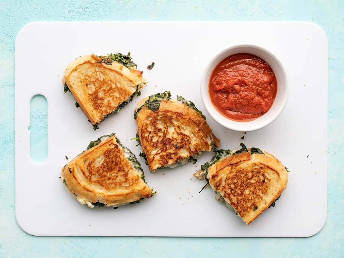Overhead view of spinach and feta grilled cheese on a cutting board with a bowl of sauce