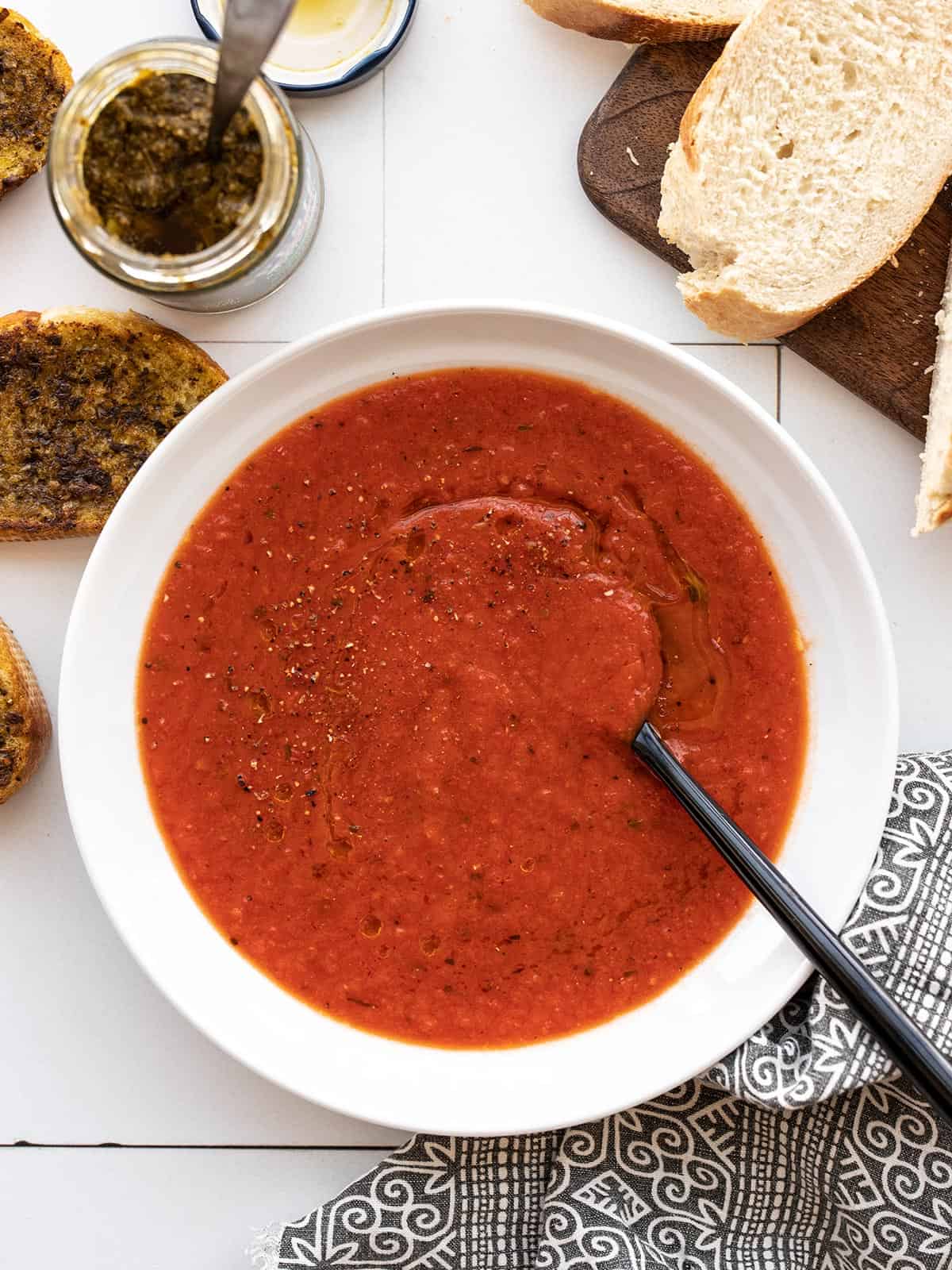 Overhead view of a bowl of roasted red pepper soup with bread and pesto on the sides
