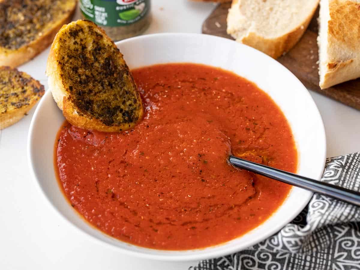 A piece of pesto toast dipped into a bowl of tomato and roasted red pepper soup
