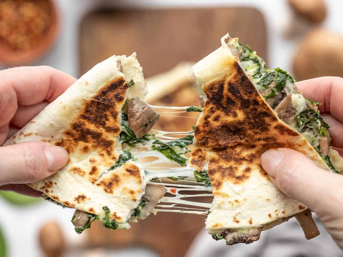 A cheesy spinach quesadilla being torn in half, cheese stretching between pieces
