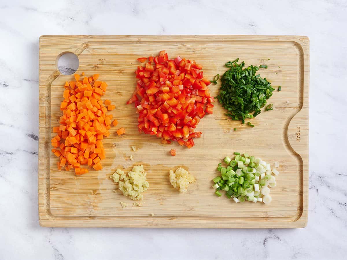 Diced carrot, bell pepper, green onions and minced garlic and ginger on a wooden cutting board.