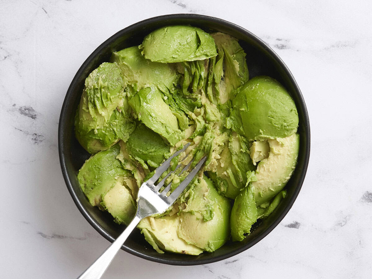 A fork mashing avocado halves in a bowl.