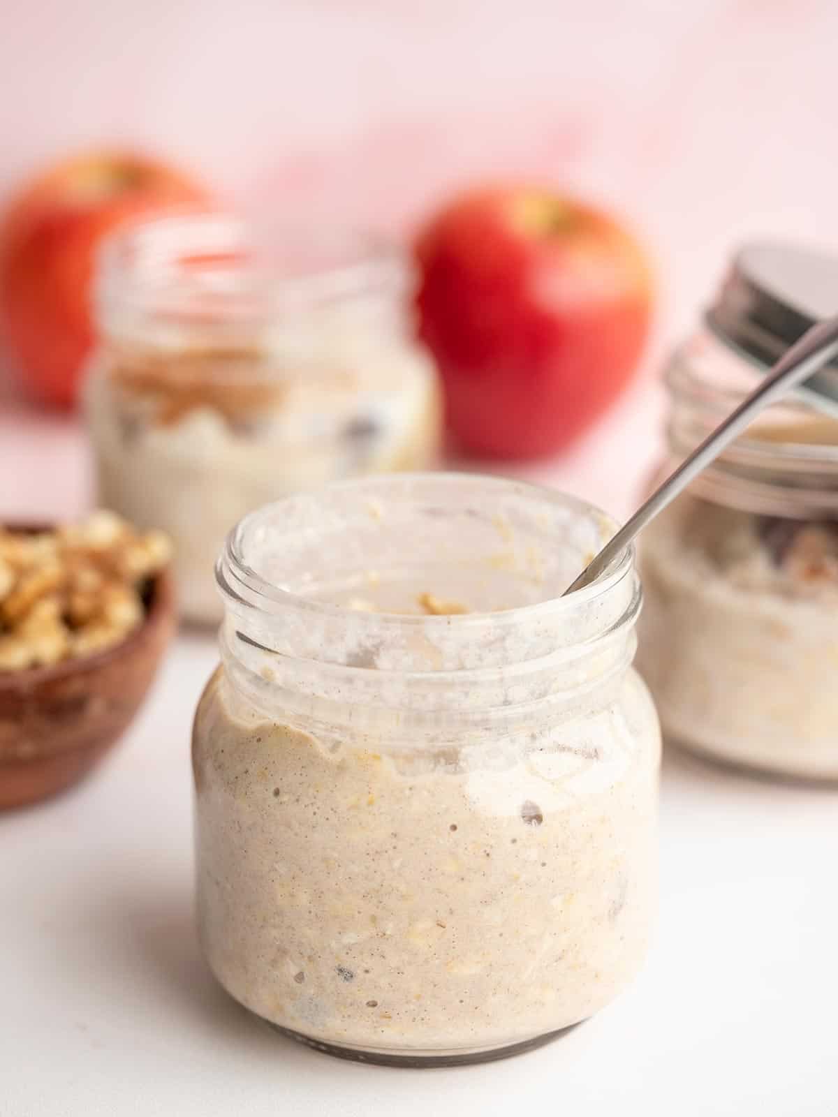 A jar of apple pie overnight oats with apples and nuts in the background.