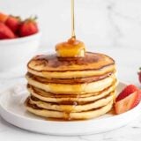 Side view of a stack of pancakes with butter on top and syrup being poured over the top.