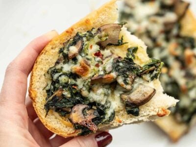 Close up of a piece of a spinach mushroom french bread pizza held in a hand.