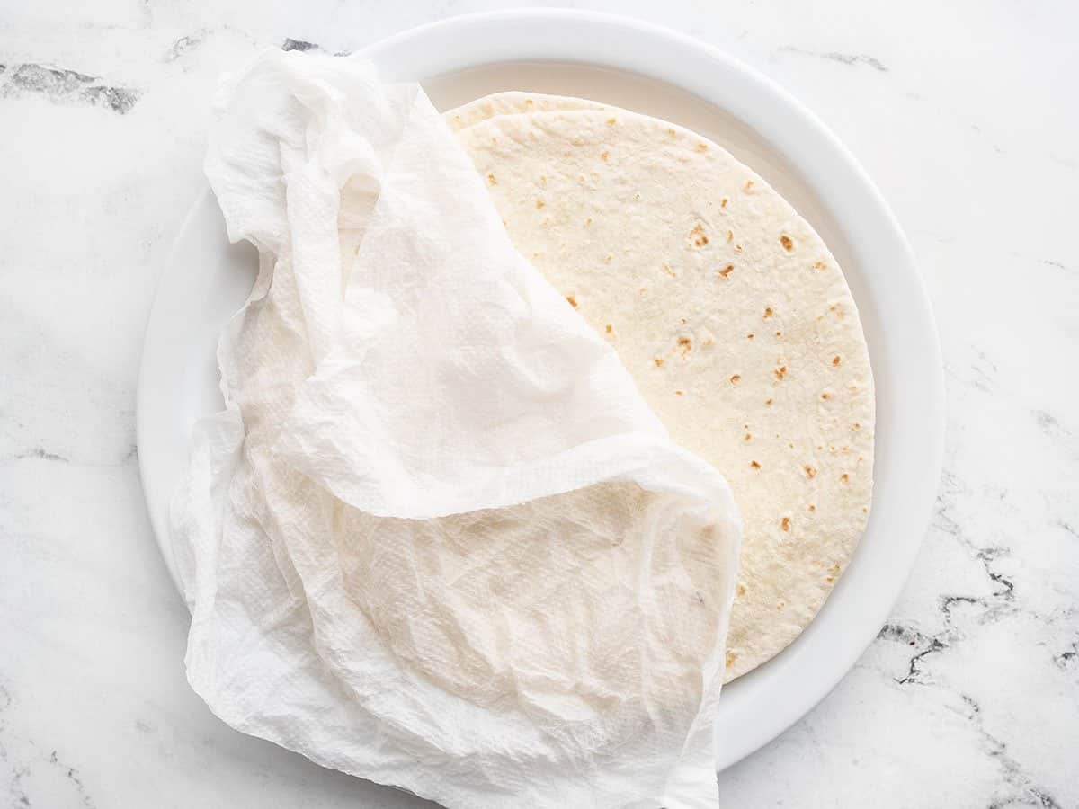Tortillas stacked on a plate with a damp paper towel covering.