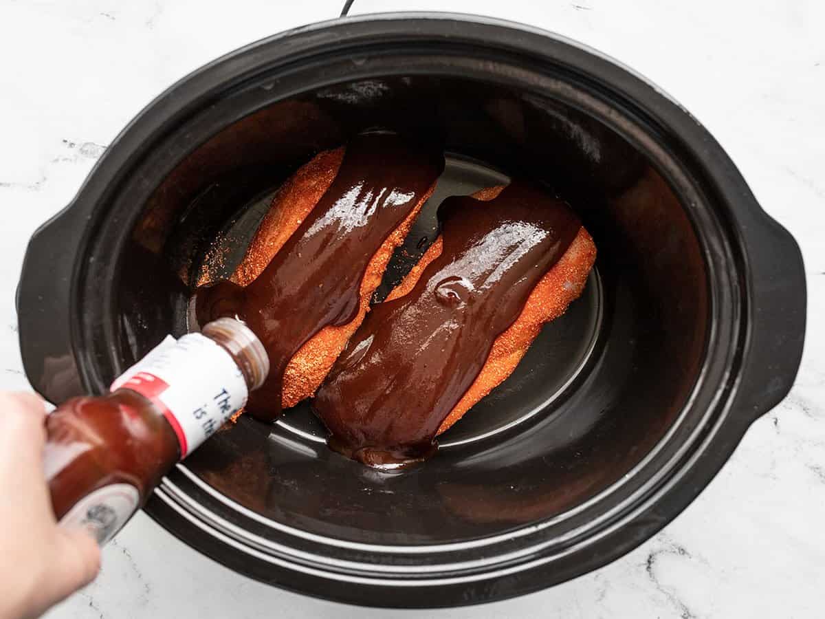 BBQ sauce being poured over chicken in the slow cooker.