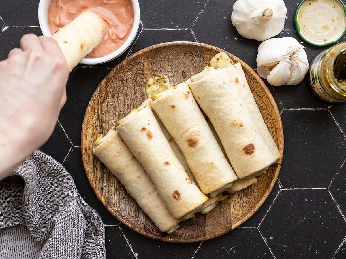 pesto mozzarella roll ups stacked on a plate, one being dipped into the sauce on the side.