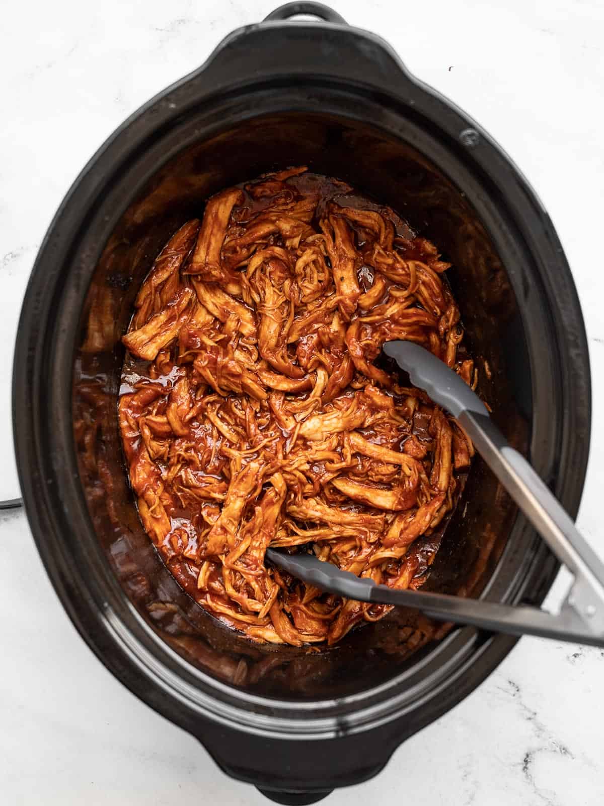 Overhead view of shredded BBQ chicken in a slow cooker with tongs.