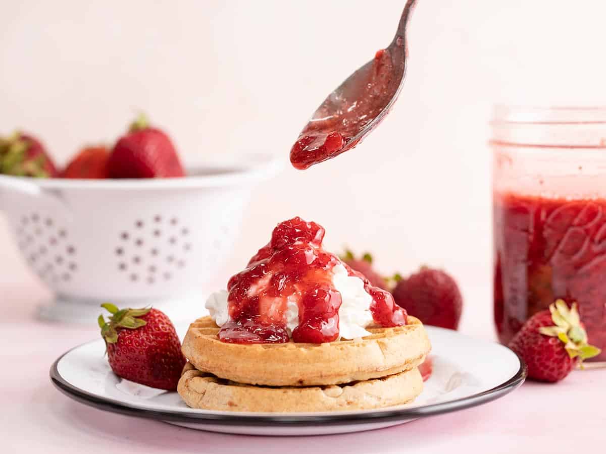 Strawberry syrup being spooned over waffles with whipped cream.