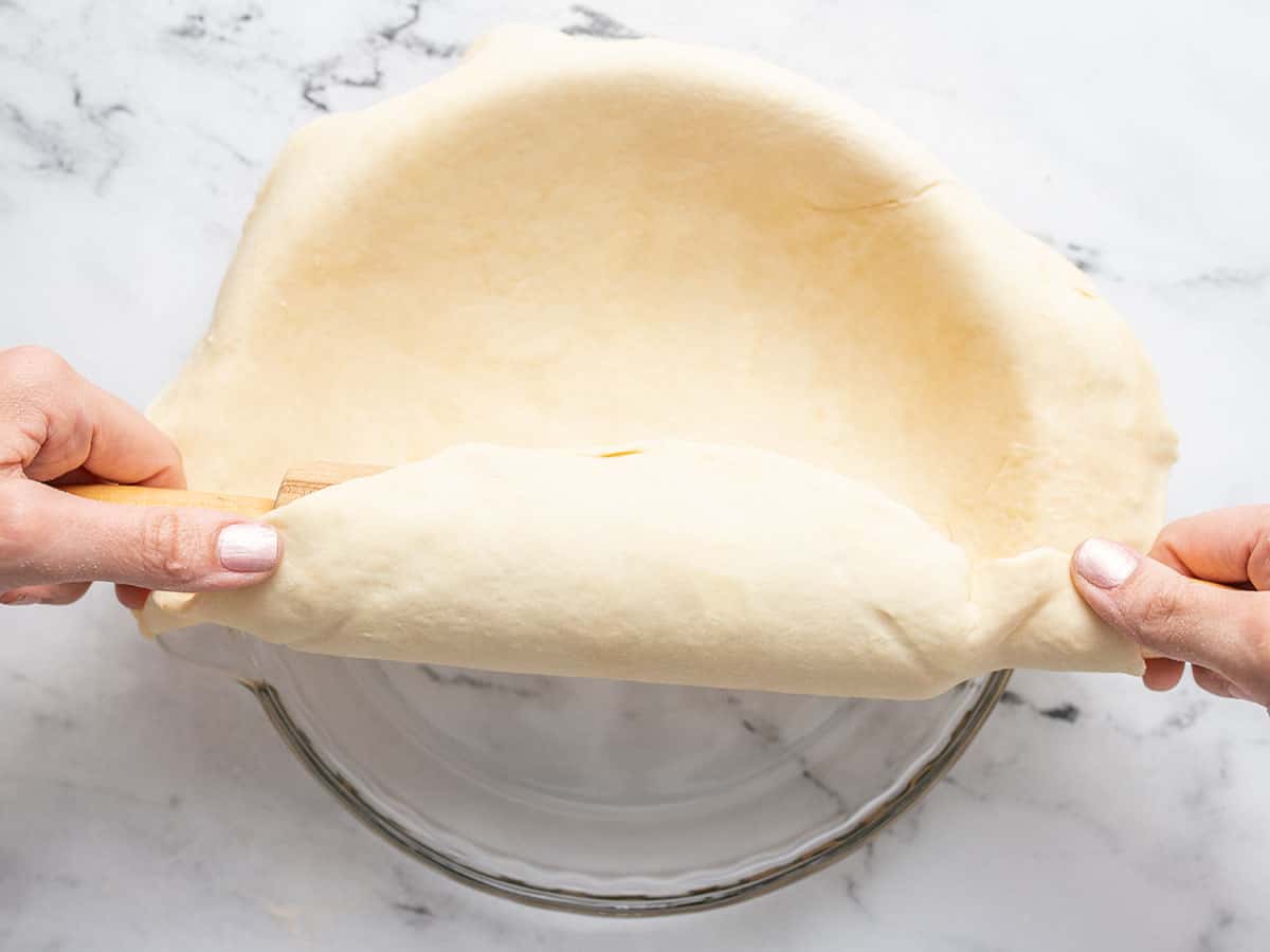 Overhead shot of two hands using a rolling pin to place rolled out dough in a pie pan.