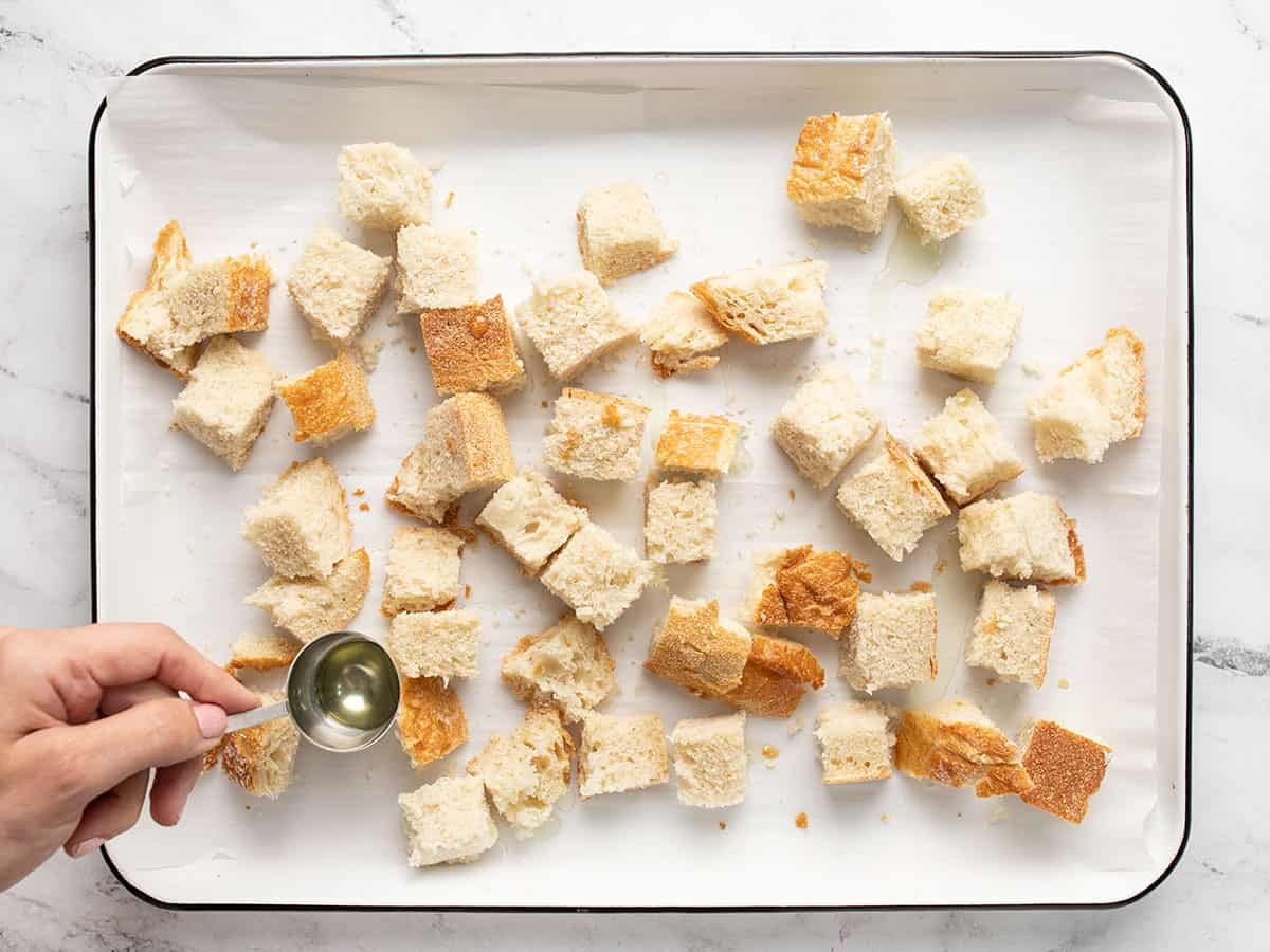 Overhead shot of cubed bread in a sheet pan being dressed with olive oil .