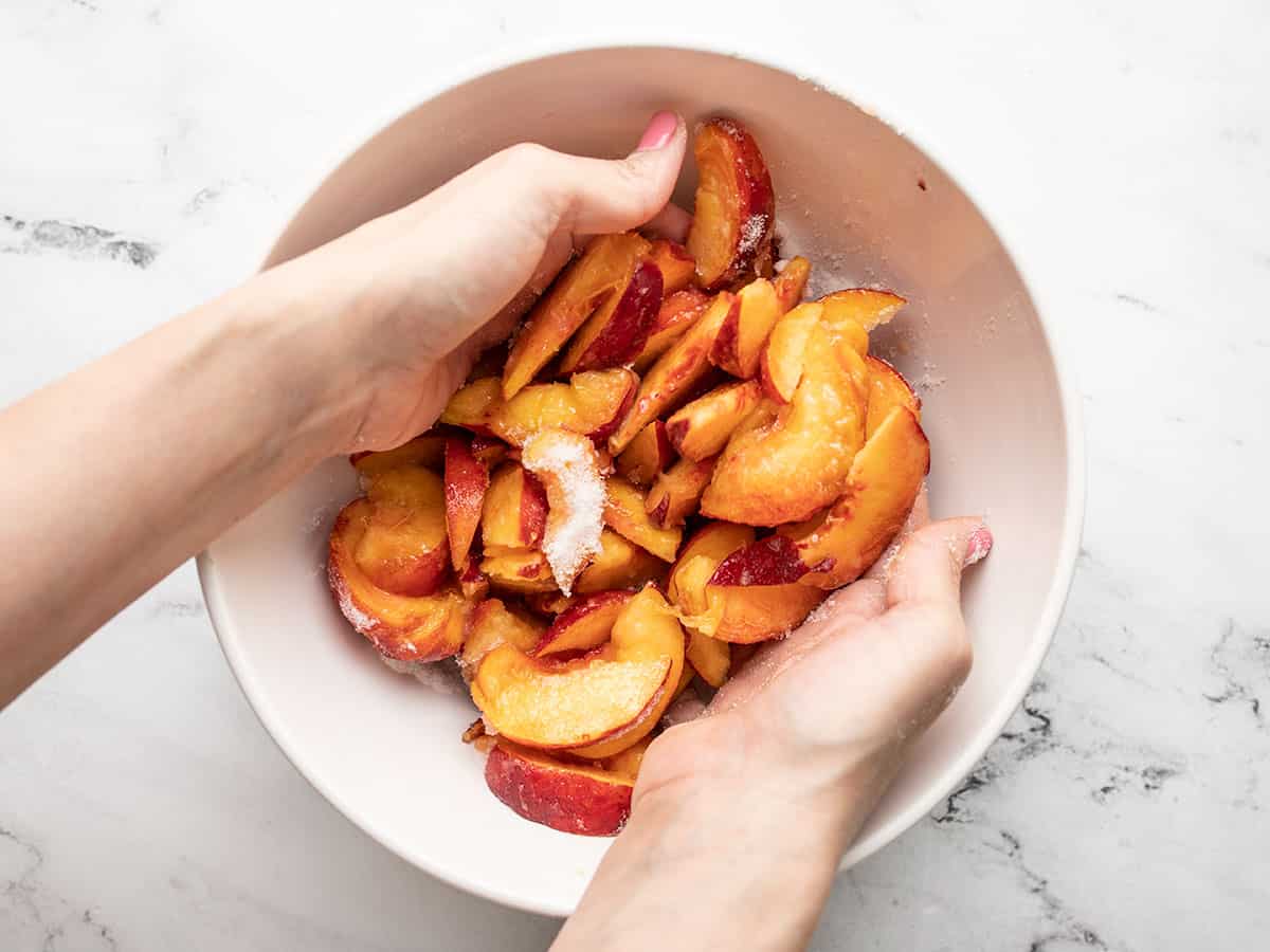 Overhead shot of wo hands tossing peaches with sugar mixture in a white bowl.