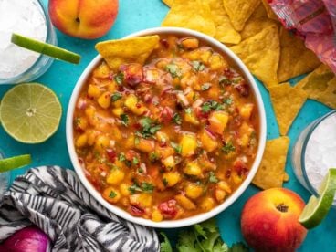 Overhead view of a bowl full of peach salsa surrounded by chips, peaches, and limes.