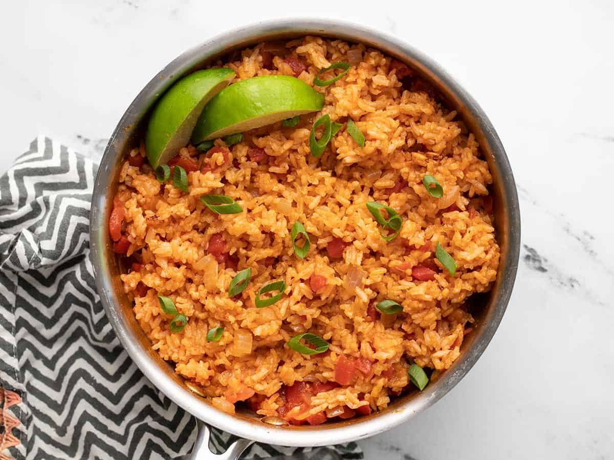 Overhead view of a pot full of tomato rice with limes and green onion.