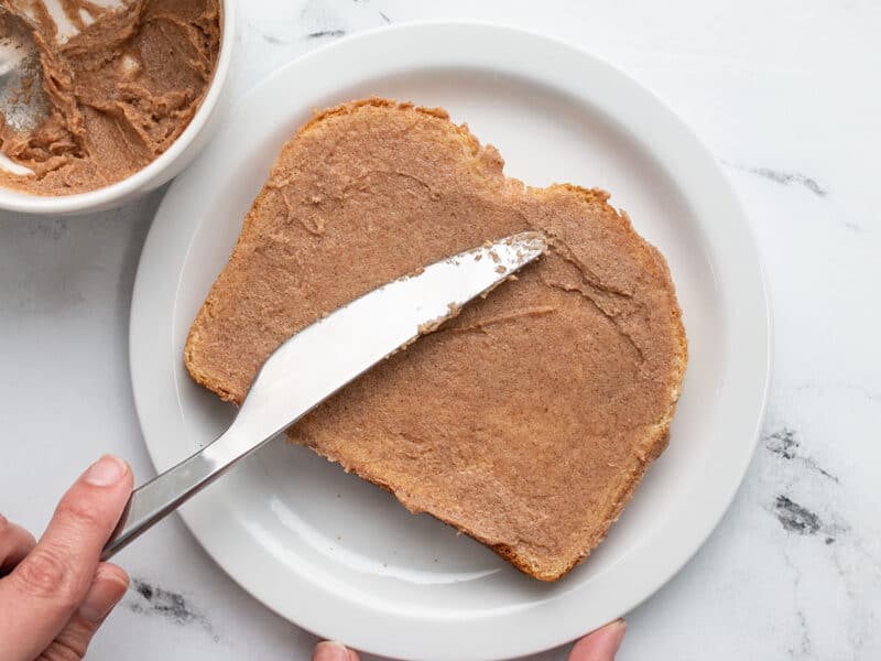 Knife spreading cinnamon butter on slice of bread on a white plate.