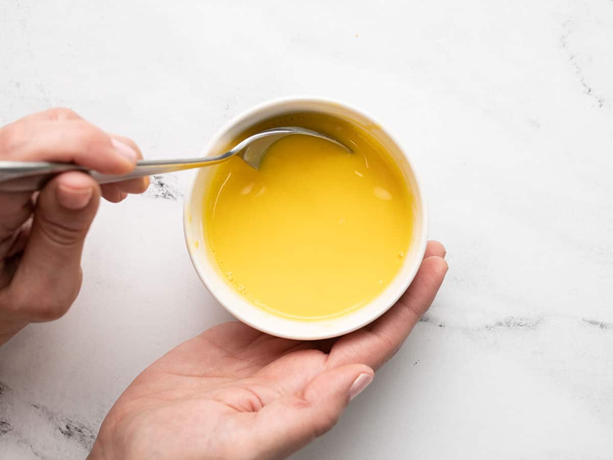 Overhead shot of egg yolks being tempered in a white ramekin with a spoon stirring it.