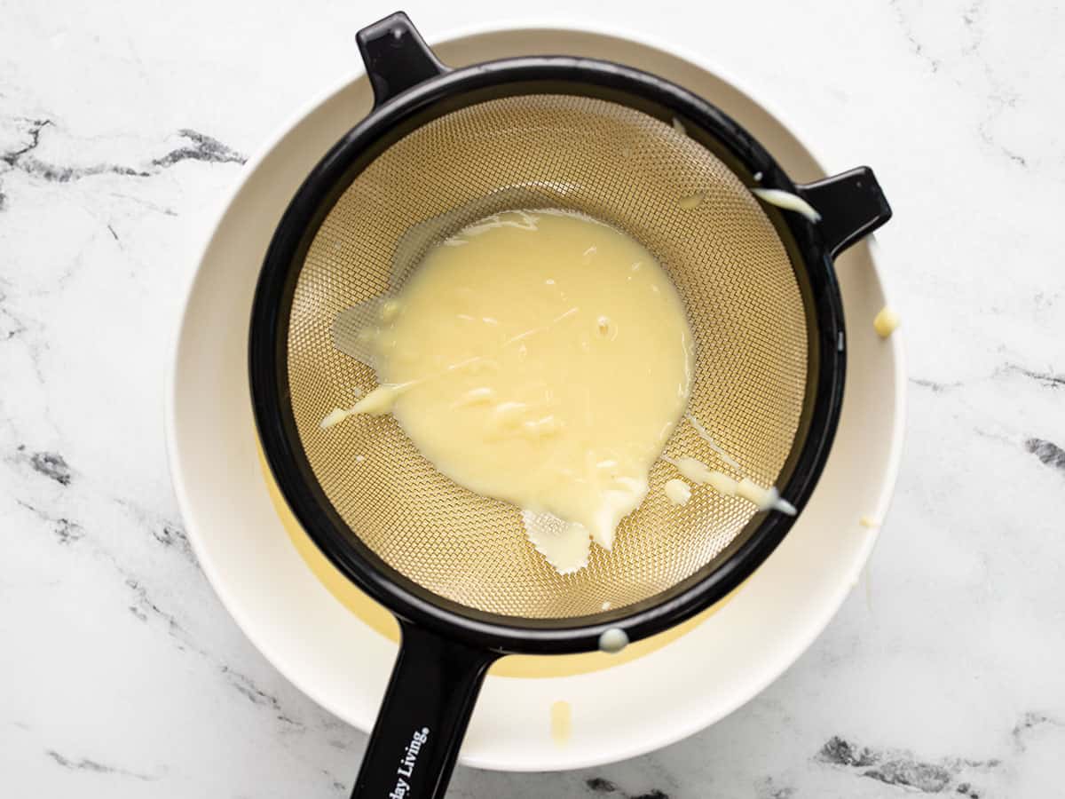 Overhead shot of vanilla pudding being strained through a fine mesh sieve into a white bowl.