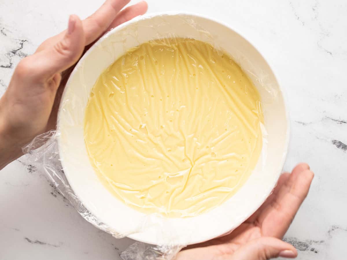 Overhead shot of vanilla pudding in a white bowl with plastic film on top.