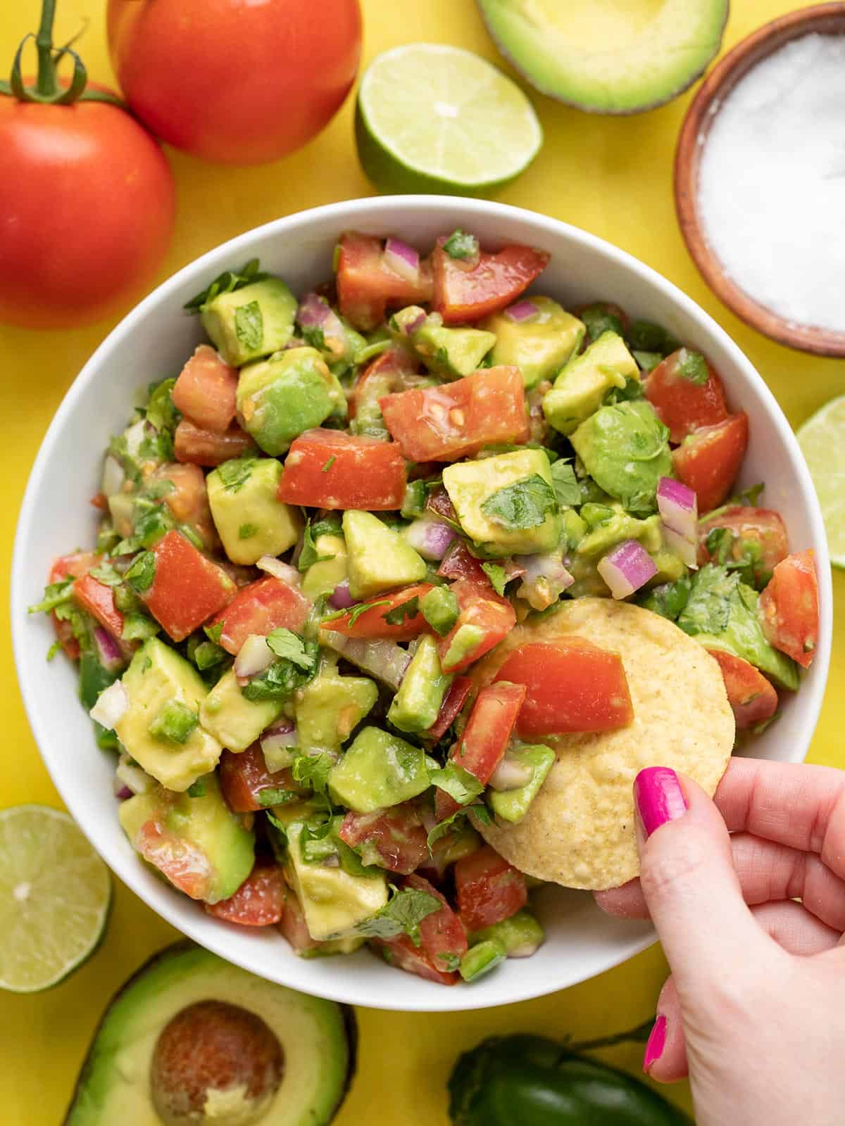 A chip dipping into a bowl of avocado and tomato salad on a yellow background.