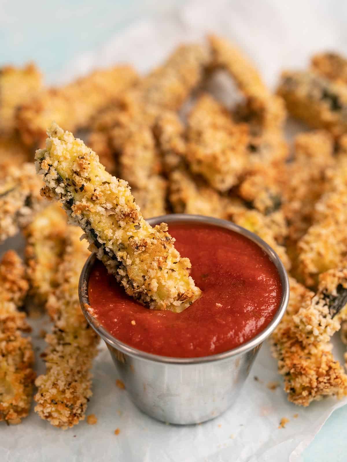 Close up of a baked zucchini fry being dipped into a cup of pizza sauce.
