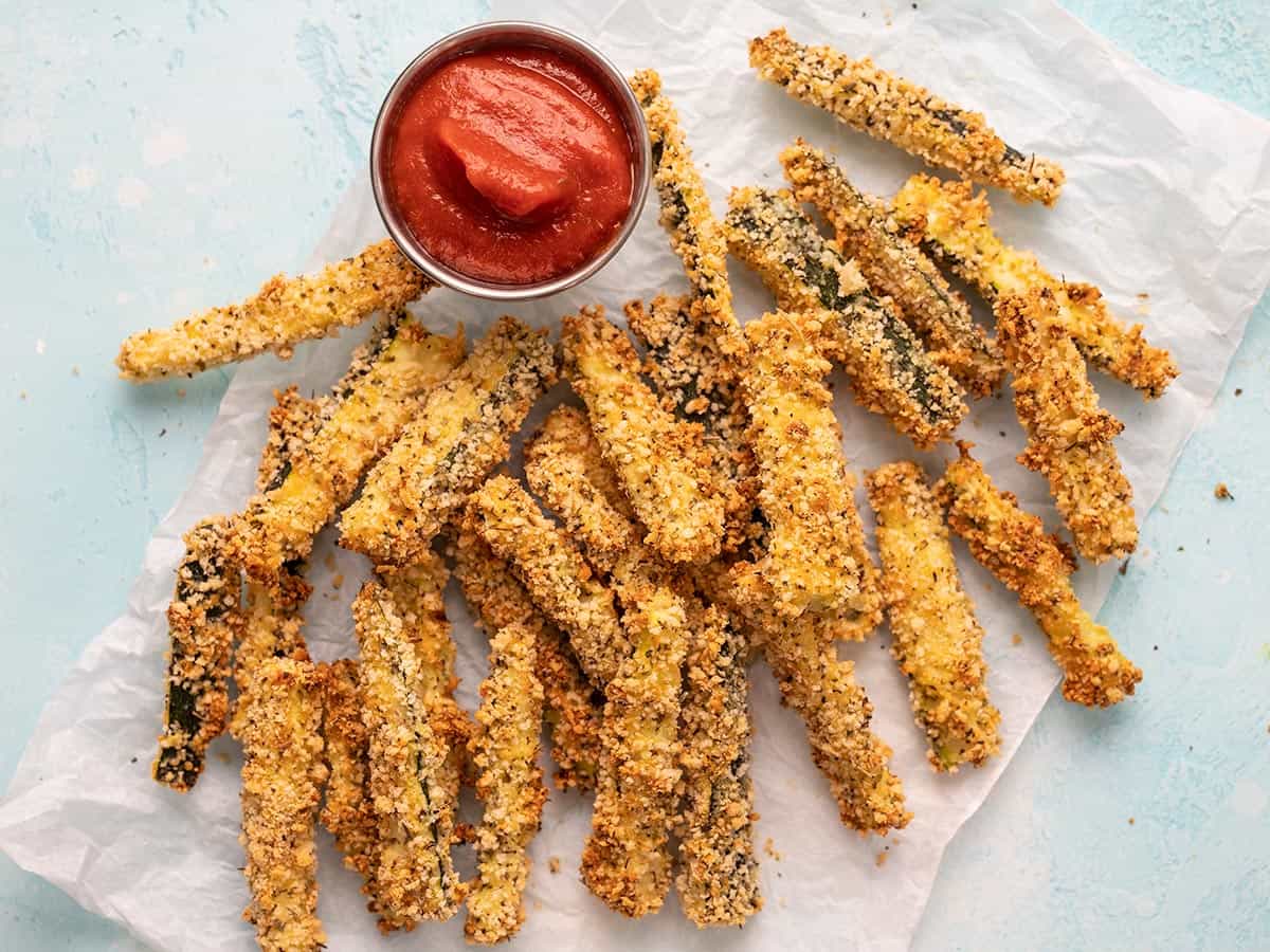 Baked Zucchini Fries on a piece of parchment with a dish of pizza sauce.