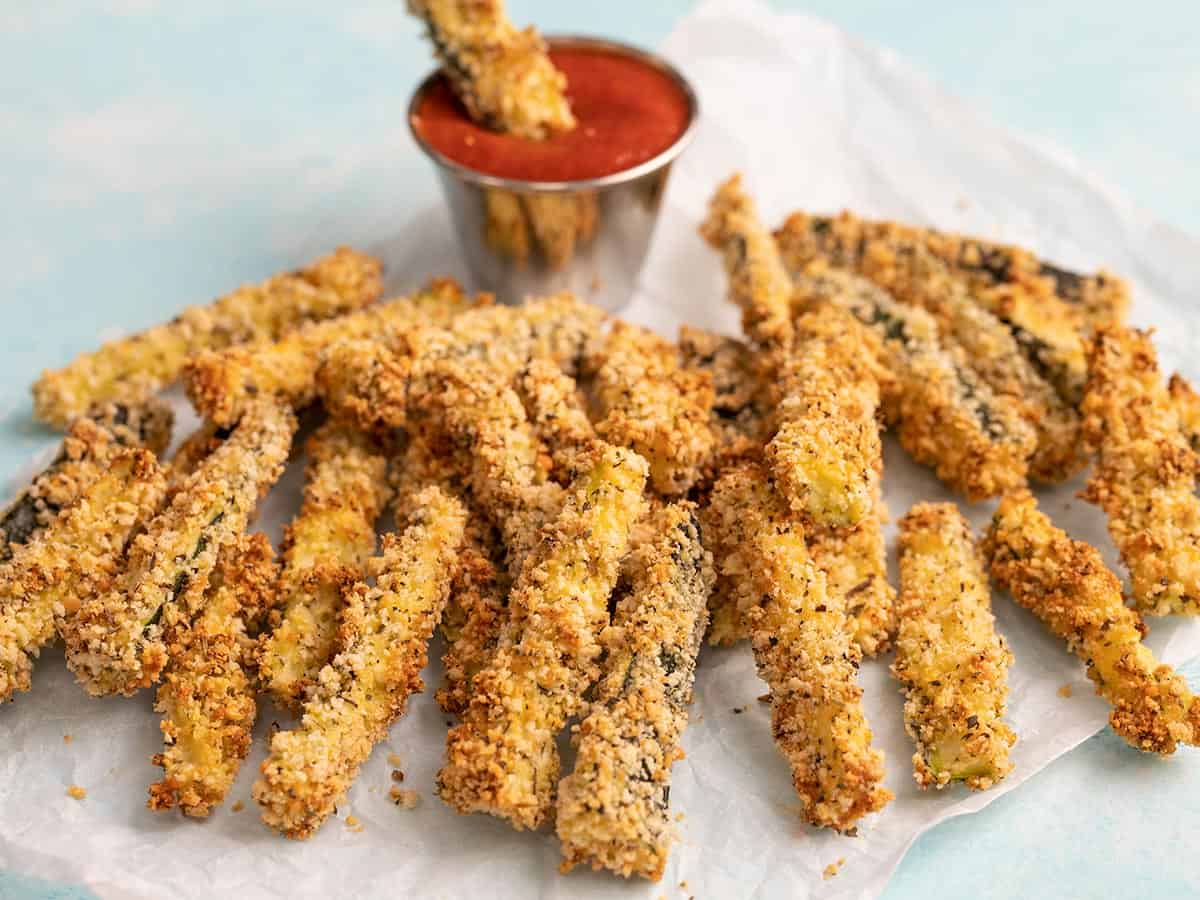 Baked zucchini fries on parchment with a dish of pizza sauce in the back.