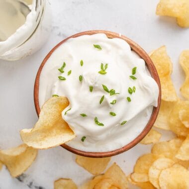 Overhead shot of a wooden bowl with sour cream in it that's topped with chives and is surrounded by potato chips.