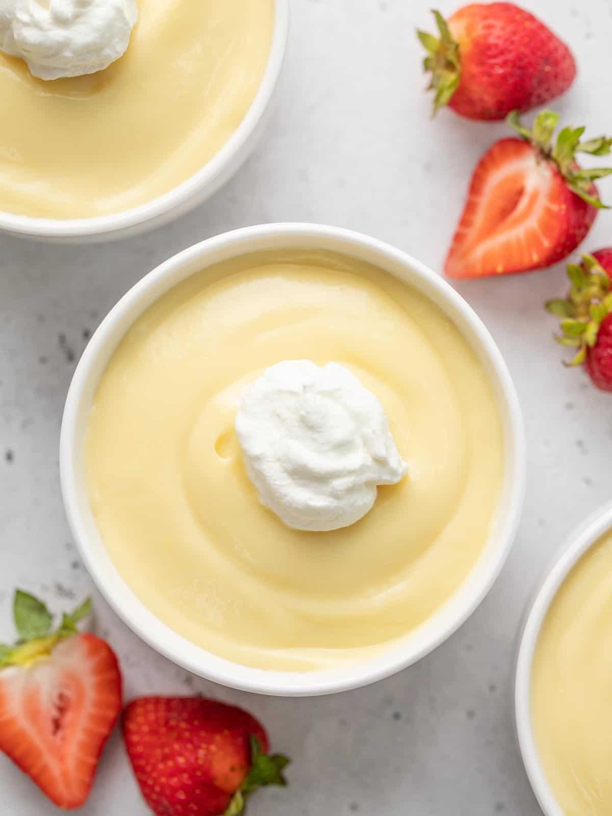 Overhead shot of vanilla pudding in a white bowl with whipped cream on top and cut strawberries on the counter.