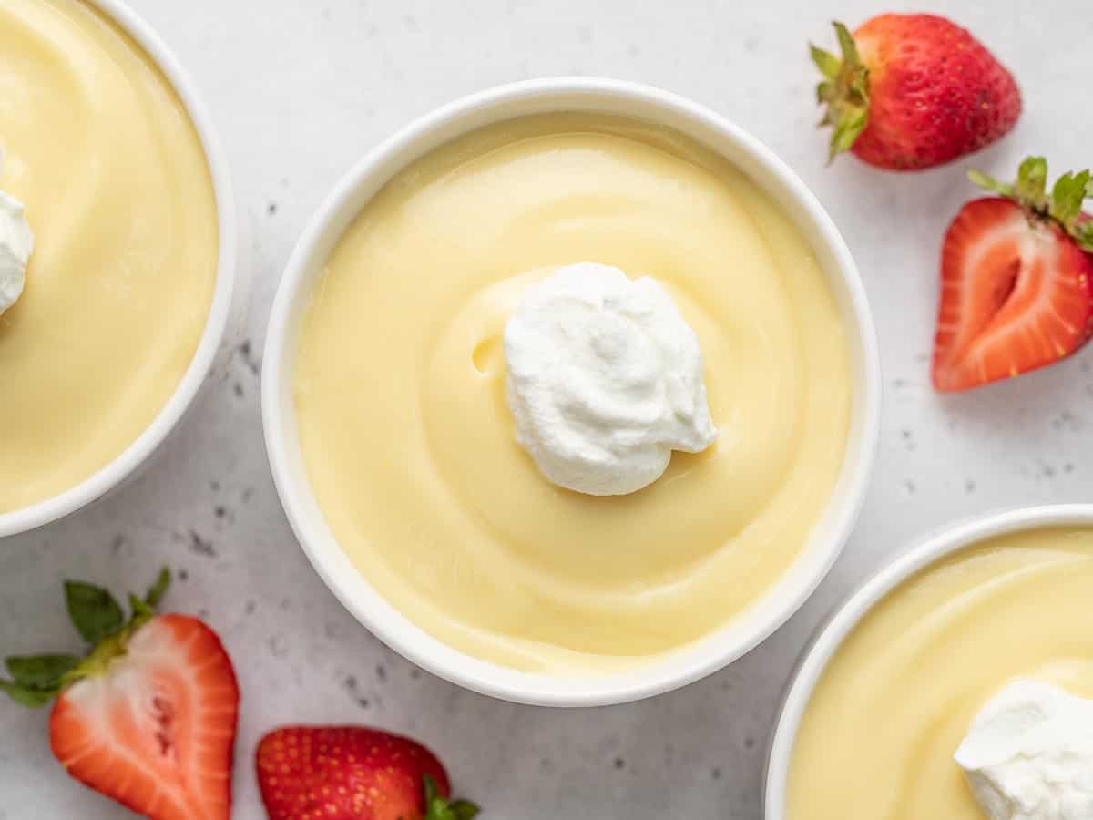 A shot of vanilla pudding in a white bowl, topped with a dollop of whipped cream and surrounded by strawberries.