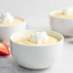 Side shot of white bowl of vanilla pudding with two other bowls and cut strawberries in the background.