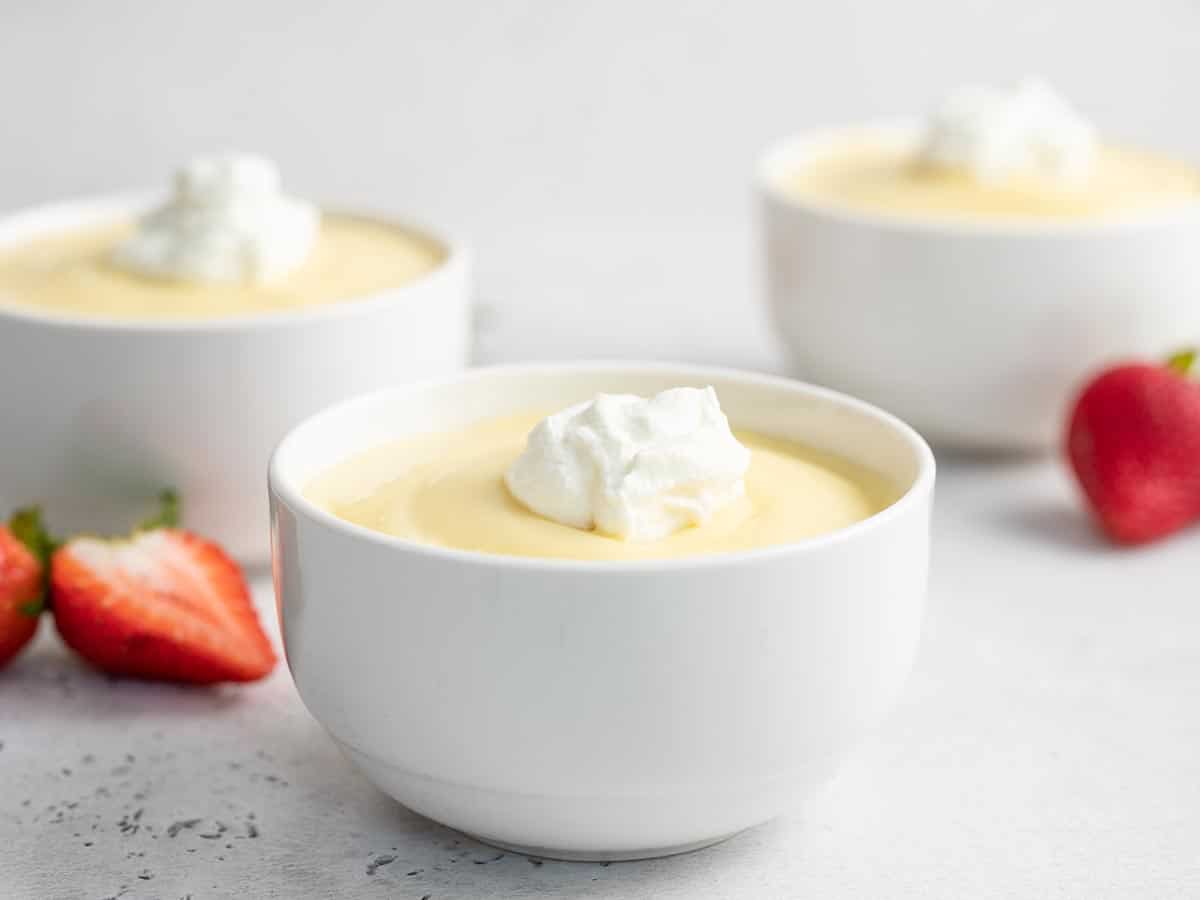 Side shot of white bowl of vanilla pudding with two other bowls and cut strawberries in the background.