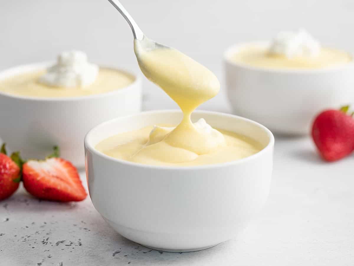 Side view of white bowl of vanilla pudding with a spoon coming out of it with strawberries and two other bowls of pudding in the background.