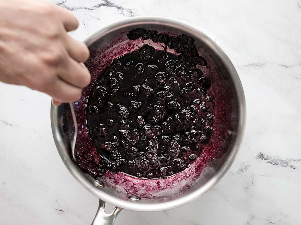 Overhead shot of hand holding a spoon and mashing blueberries in a pot.