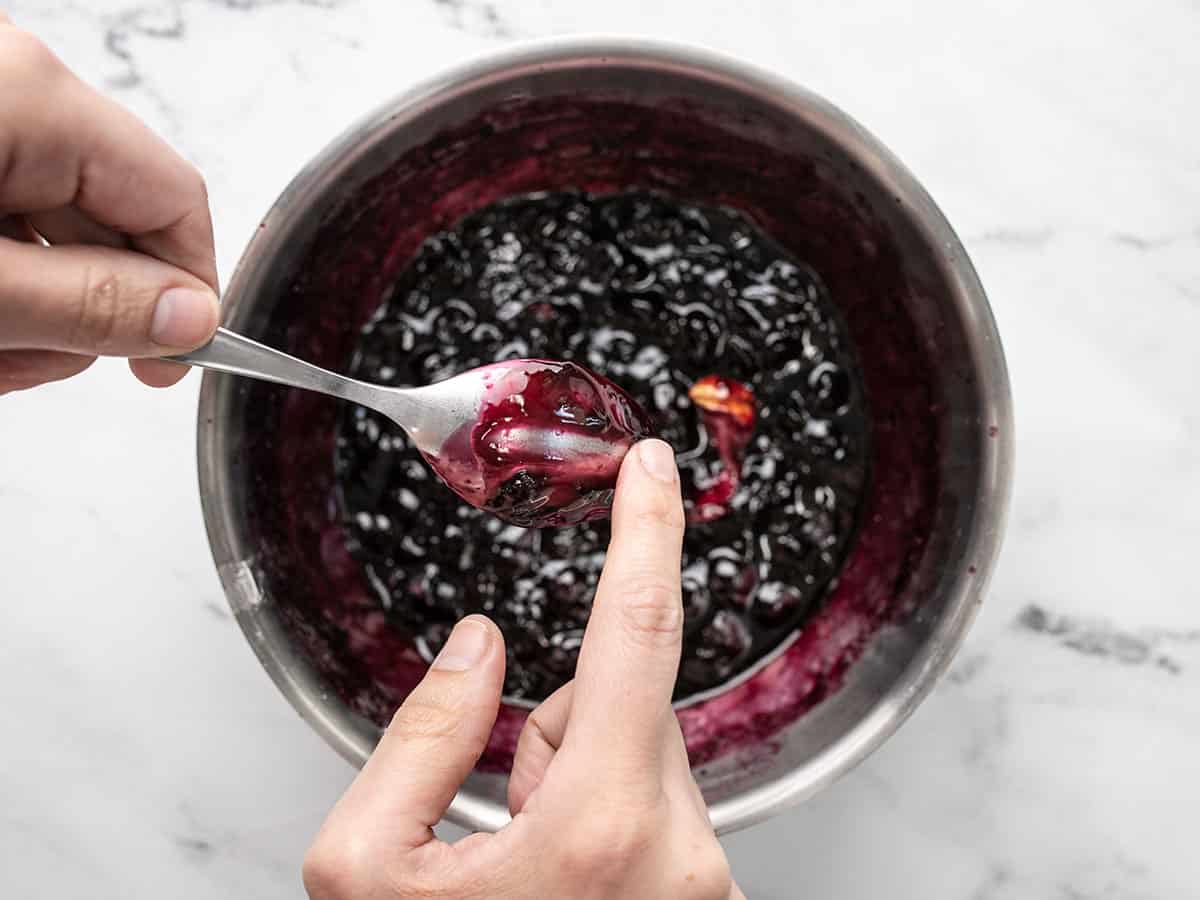 Overhead shot of blueberry sauce in a pan with finger checking sauce thickness on the back of a spoon.