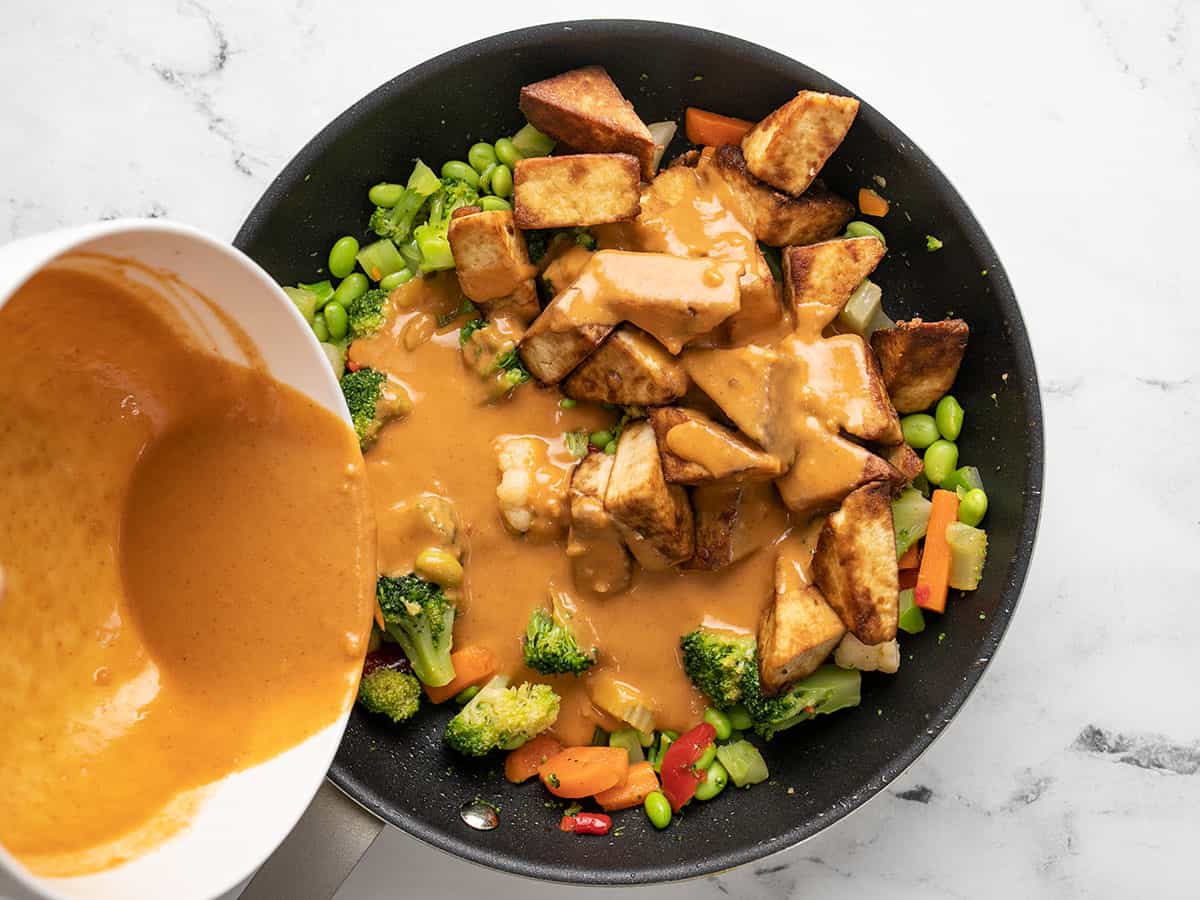 Tofu and peanut sauce being added to the skillet with vegetables.