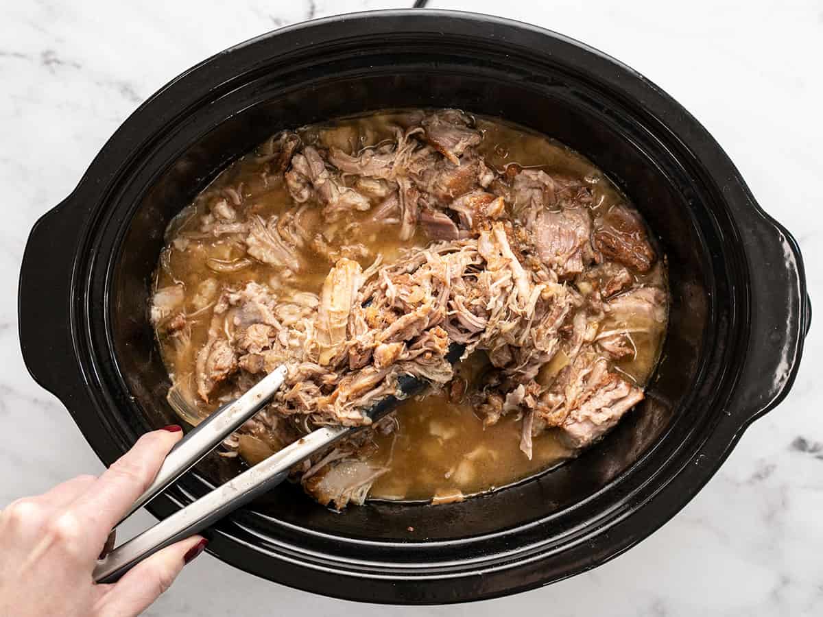 Shredded pork in the slow cooker, some being lifted with tongs.