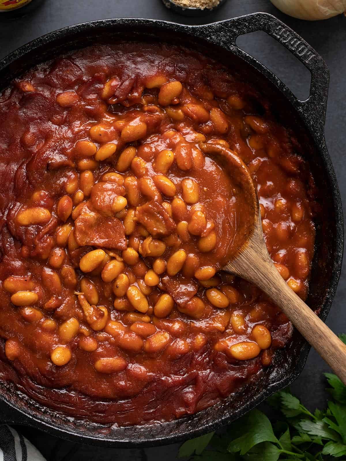 Close up overhead view of baked beans in a cast iron skillet with a wooden spoon.