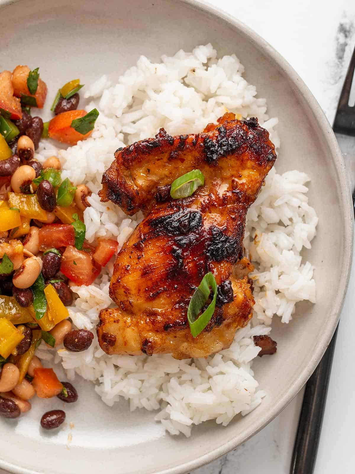 A honey chipotle chicken thigh on a bed of rice on a plate next to bean salad.