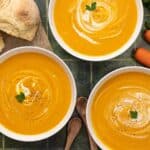 Overhead shot of carrot soup in three white bowls with sour cream swirled in it