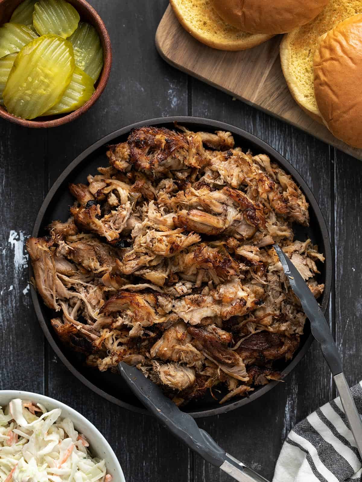Slow cooker pulled pork on a plate with tongs and sandwich fixings on the sides.